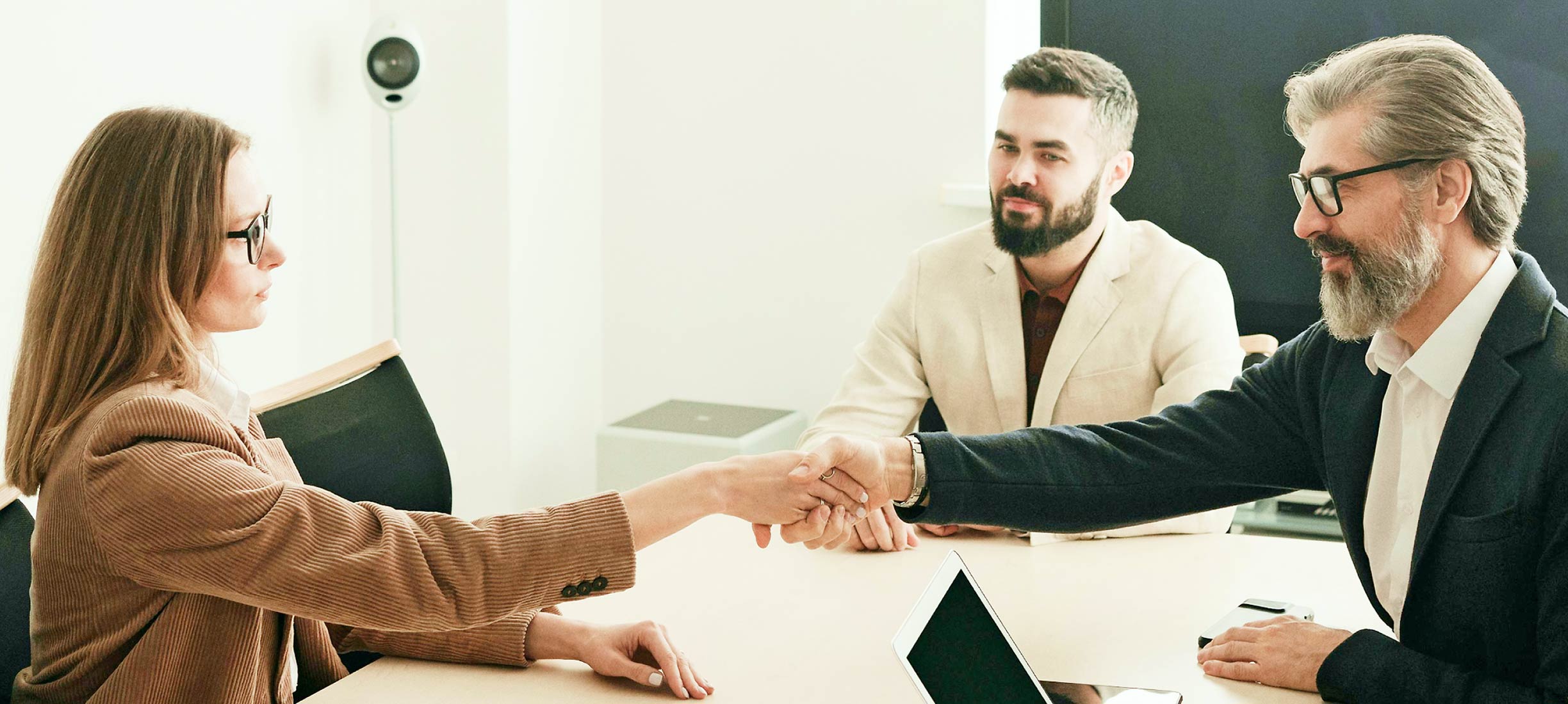 Zwei Personen reichen sich bei einem Meeting über den Tisch die Hand, während eine dritte Person daneben sitzt.