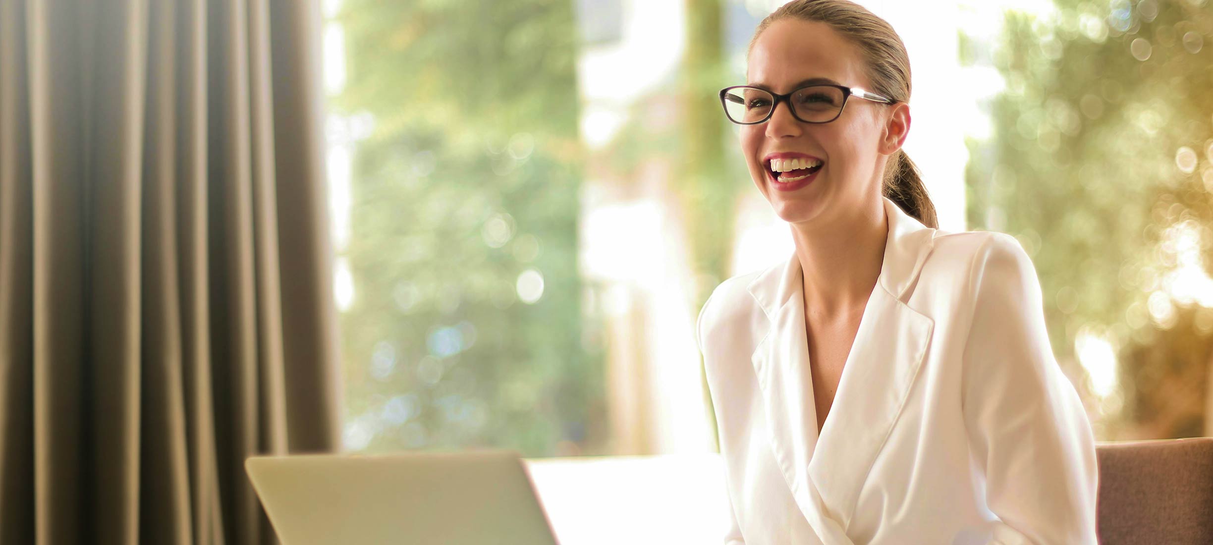 Frau mit Brille arbeitet lächelnd an einem Laptop in einem hellen Büro.