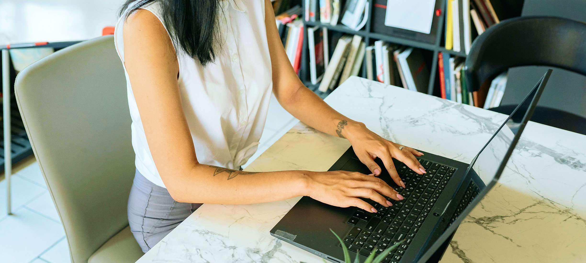 Person tippt auf einem Laptop an einem Schreibtisch mit Unterlagen.