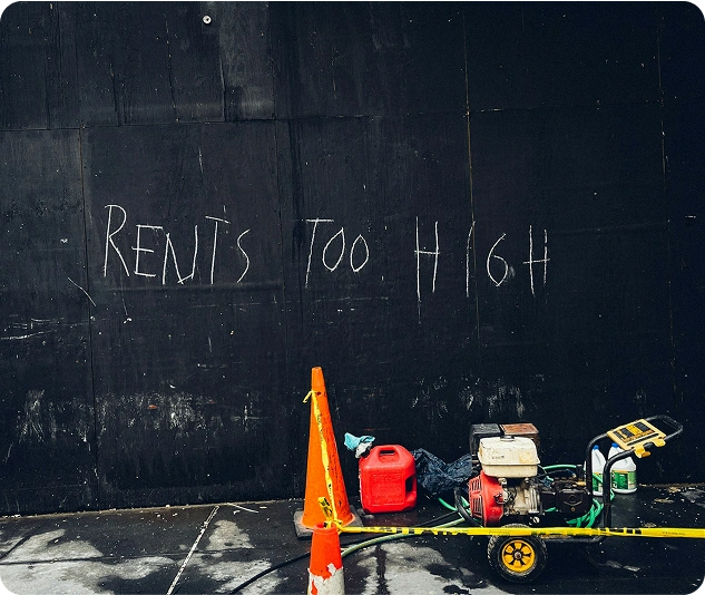 Graffiti mit der Aufschrift „Rent’s Too High“ auf einer dunklen Wand, davor Baugeräte und ein Warnkegel.