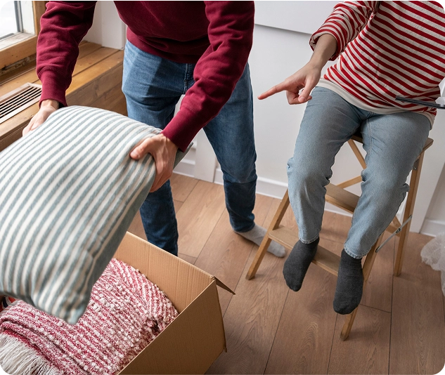 Person packt Kissen in einen Umzugskarton, während eine andere Person daneben sitzt und darauf zeigt.