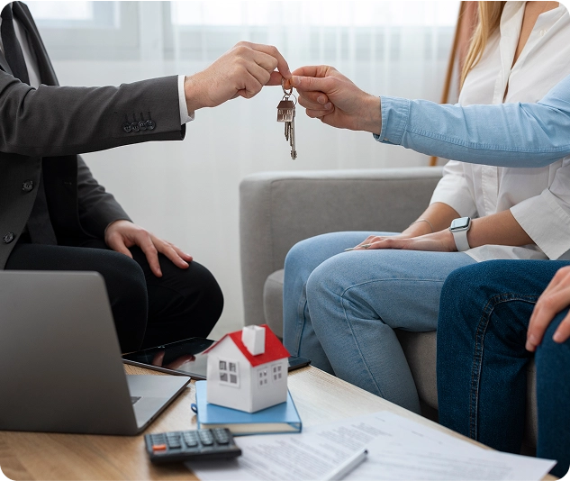 Person übergibt einem Paar einen Schlüssel während eines Treffens mit Unterlagen, Laptop und einem Modellhaus auf dem Tisch.