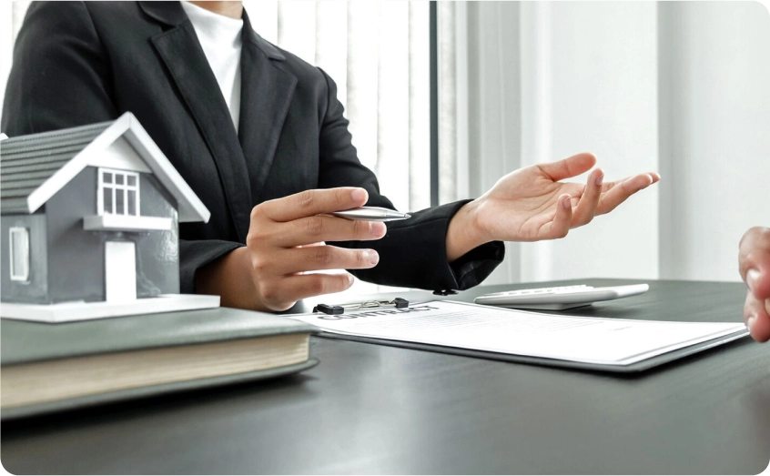 Person erklärt etwas über ein Dokument, neben einem Modellhaus auf dem Tisch.