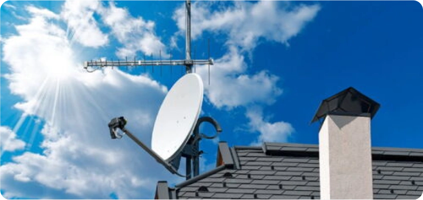 Satellitenschüssel und Antenne auf einem Hausdach unter blauem Himmel.