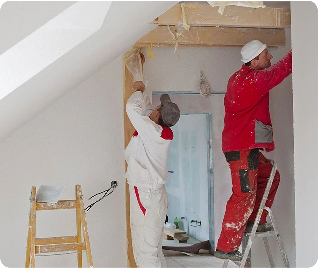 Zwei Handwerker in Arbeitskleidung renovieren einen Türdurchgang, einer steht auf einer Leiter und arbeitet an der Wand.