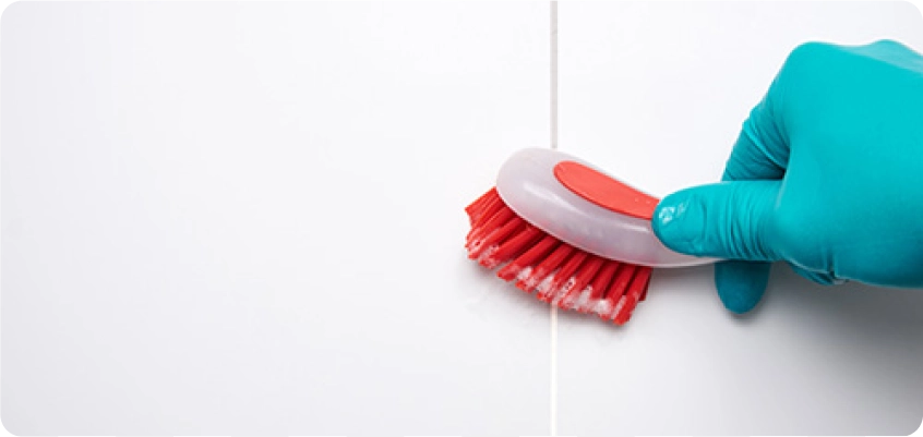 Hand mit Reinigungshandschuh schrubbt eine Fliesenfuge mit einer Bürste.